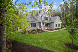 A charming gray two-story house surrounded by lush green lawn, trees, and well-maintained landscaping under a clear sky.