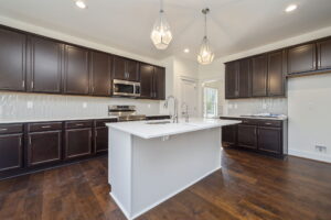 A modern kitchen features dark wood cabinets, a white central island with a sink, stainless steel appliances, pendant lights, and wood flooring.