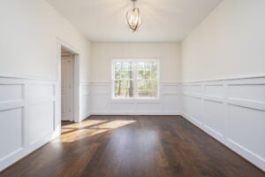 A bright, empty room with white wainscoting, hardwood floors, a central light fixture, and a window showing outdoor greenery. There's an open door to the left side.