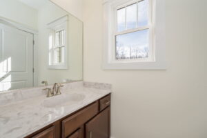 A bathroom with a marble countertop, single faucet sink, and a window with a view of trees outside. White walls and a large mirror above the sink.