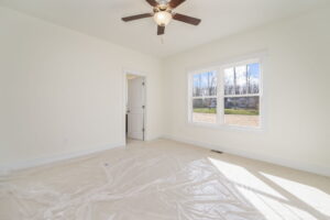 A sparsely furnished room with cream-colored walls, a ceiling fan, a window with a view of trees, and plastic sheeting covering the light carpeted floor. A white door is partially open.