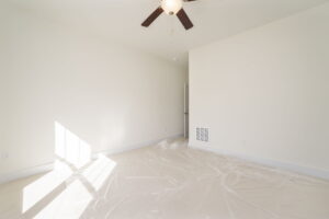 An empty room with white walls and carpet, a ceiling fan, plastic sheets on the floor, and sunlight streaming through a window.