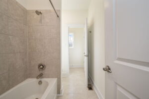A tiled bathroom with a bathtub and showerhead on the left. An open door leads to a corridor with beige flooring and white walls. Another door is partially open on the right.