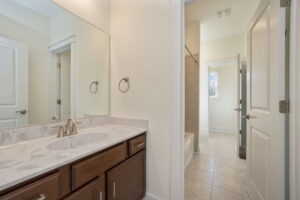 Modern bathroom with a marble countertop, wood cabinets, and a large mirror. The view extends to a hallway with tiled flooring, a closed door, and a bathtub area with a shower.