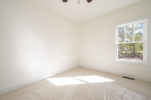 An empty, sunny room with beige walls, a window on the right, and a ceiling fan. The floor is covered with protective plastic sheeting.