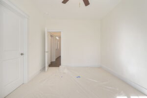 A bright, empty room with a plastic sheet covering part of the carpeted floor. The room has a white door that's open, revealing a hallway. A ceiling fan with dark blades is visible.