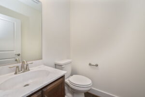 A small bathroom with a toilet, a sink with a marble countertop, and a large mirror. An empty toilet paper holder is mounted on the wall beside the toilet.