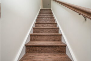 A set of wooden stairs with a handrail on the right side, leading up between two white walls.
