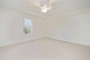 An empty room with beige carpet, off-white walls, and a ceiling fan with a light. There is a window on one wall allowing natural light in.
