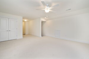 An empty carpeted room with white walls, a ceiling fan, double doors on the left, a small closet, and a light fixture in the hallway.