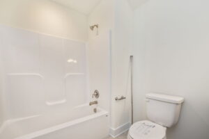 A bathroom features a white bathtub with a showerhead, a silver faucet, and a white toilet. There is a silver grab bar beside the toilet. The walls are white.