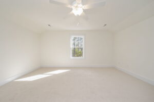 A spacious, empty room with light beige carpet, white walls, a ceiling fan with a light, and a single window letting in natural light.