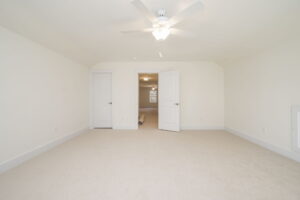 An empty room with beige carpet, white walls, a ceiling fan, and two open doors leading to another room.