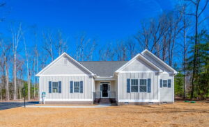 A single-story white house with a front porch, large windows, and a grey shingled roof, set in front of leafless trees under a clear blue sky.