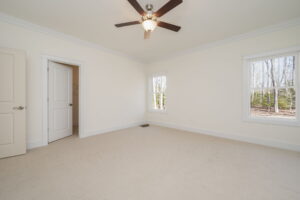 An empty carpeted room with light-colored walls, a ceiling fan, two windows showing an outdoor scene with trees, and an open door leading to a closet.