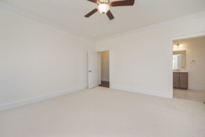 Empty carpeted bedroom with a ceiling fan, beige walls, an open door leading to an adjacent room, and a doorway to a bathroom with a vanity and mirror.