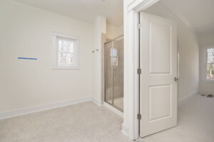 A clean, bright bathroom with a walk-in glass shower, white walls, and a window. Adjacent room has white carpet and an open door.
