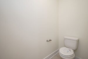 A small minimalist bathroom with a white toilet and an empty toilet paper holder on a light-colored wall. The floor is tiled with light gray tiles.