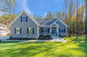 Single-story gray house with white trim and a gabled roof, featuring a front porch and surrounded by a grassy lawn with trees in the background on a sunny day.