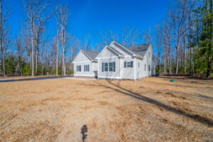 A newly constructed white house with a grey roof sits in a barren yard with scattered straw, against a backdrop of leafless trees under a clear blue sky.
