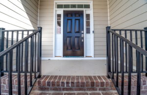 A front door painted black with two white side windows is centered between beige siding walls. Brick steps lead up to the door, framed by black metal railings.