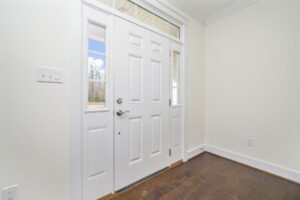 A white front door with glass panels on either side and above. The door has a metal handle and lock. The entryway has light-colored walls and dark wood flooring.