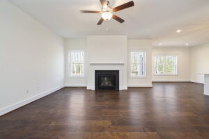 A spacious, empty living room with a ceiling fan, hardwood floors, a fireplace, and large windows. An adjoining area with similar flooring is visible to the right.