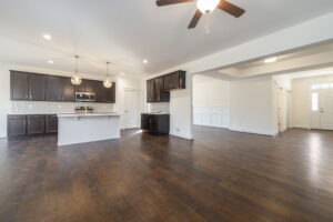 An empty, modern open-concept kitchen and living area featuring dark wood cabinets, an island, hardwood floors, and white walls under recessed and pendant lighting with a ceiling fan.