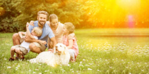 A happy family with two children and a dog sitting on grass in a sunny park, laughing together.