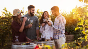Two couples enjoy wine and grapes at a vineyard during sunset, standing around a barrel, smiling and toasting glasses.