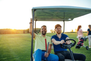Two men smiling and sitting in a golf cart at a course during sunset, with other players in the background.