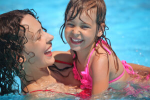 A joyful woman and young girl with wet hair laughing together in a swimming pool, both wearing swimsuits.