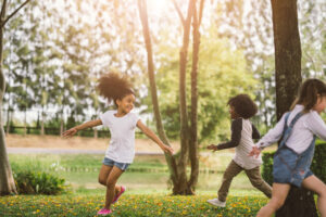 Three children playing tag in a sunlit park, with one girl in a white shirt joyfully running ahead of the others.