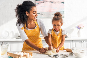 A woman and a young girl in matching yellow aprons kneading dough together in a bright kitchen.