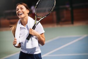 A joyful woman with a tennis racket draped over her shoulder and a towel around her neck, laughing on a tennis court at night.