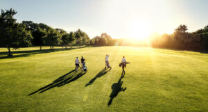 Four golfers walking on a sunny golf course with bags, casting long shadows on the grass, during sunset.