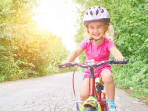A young girl wearing a helmet and smiling while riding a bicycle on a tree-lined path.