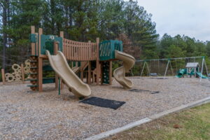A playground with two slides, climbing structures, and a swing set, surrounded by trees and covered with wood chips.