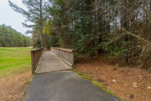 Wooden footbridge over a small dip in a park, surrounded by pine trees and a grassy area under a cloudy sky.