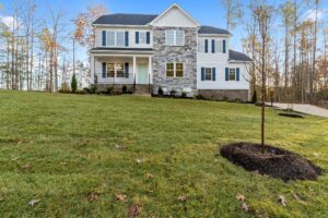 A two-story suburban home with a gray and white facade, surrounded by a freshly mowed lawn and young trees under a clear blue sky.