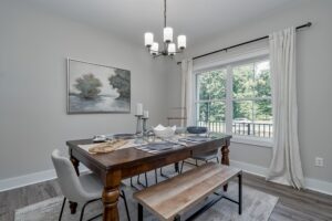 Modern dining room with a wooden table, benches, a chandelier, and a landscape painting, overlooking a window with a view of greenery.