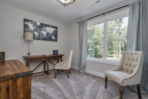 A home office with a wooden desk, beige upholstered chair, chest drawer, and framed artwork on the wall next to a window with gray curtains.