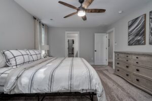 A modern bedroom featuring a neatly made bed with striped bedding, a wooden dresser, gray carpet, and a ceiling fan, with doors leading to other rooms.