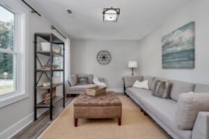 Modern living room with gray sofas, a brown ottoman, white walls, a black bookshelf, and wall art.