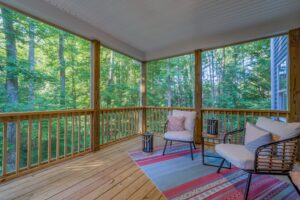 A cozy covered porch with wooden flooring, wicker chairs, cushions, and a patterned rug, surrounded by lush green trees.