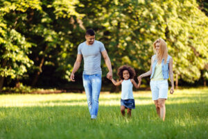 A happy family, a man, woman, and young child, walking hand in hand and smiling through a sunny park with lush green trees in the background.