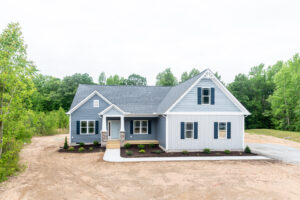 New gray single-story house with white trim and a gabled roof, surrounded by young trees and a gravel driveway.