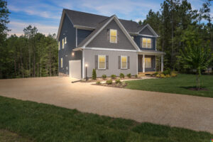 A two-story house with gray siding, white trim, and a spacious driveway, surrounded by a grassy lawn and trees in the background, illuminated by outdoor lights during the early evening.