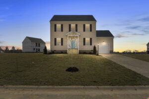 A two-story house with a manicured lawn at dusk, featuring a small tree planted in the front yard and an adjacent garage with its light on. Another house is under construction in the background.