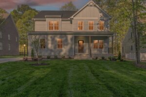 Two-story gray house with a front porch, illuminated windows, and a well-maintained lawn, surrounded by trees and neighboring houses under a dusky sky.